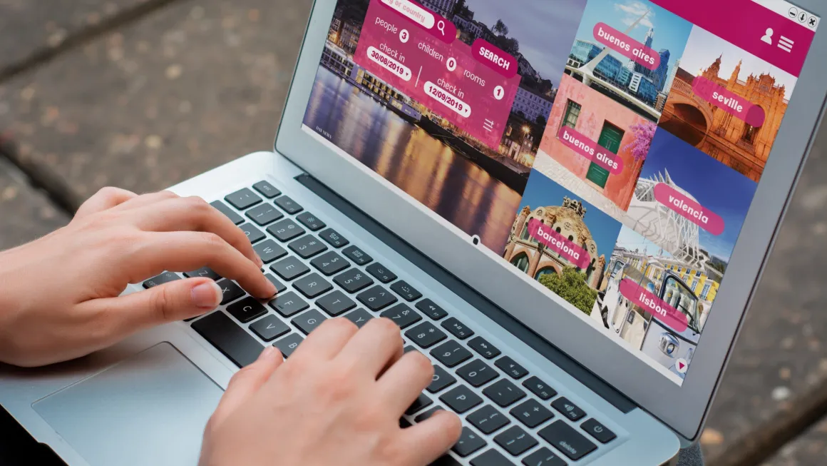 Manos tecleando en una laptop con una página web de búsqueda de alojamiento y un mosaico de ciudades en pantalla